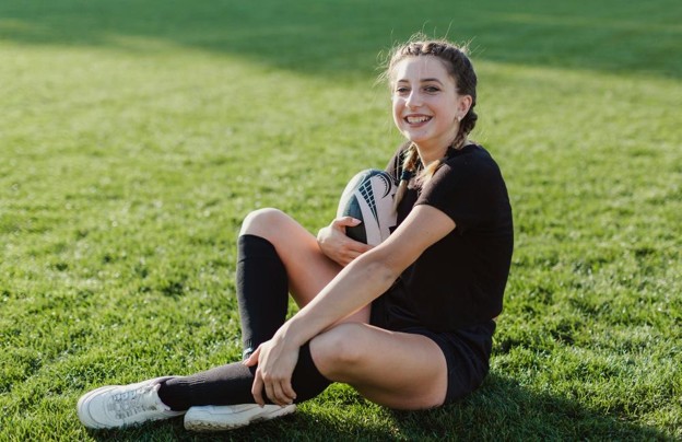 A smiling female athlete sitting on a grass field holding a rugby ball, showcasing a positive outcome of identifying common signs of overtraining.