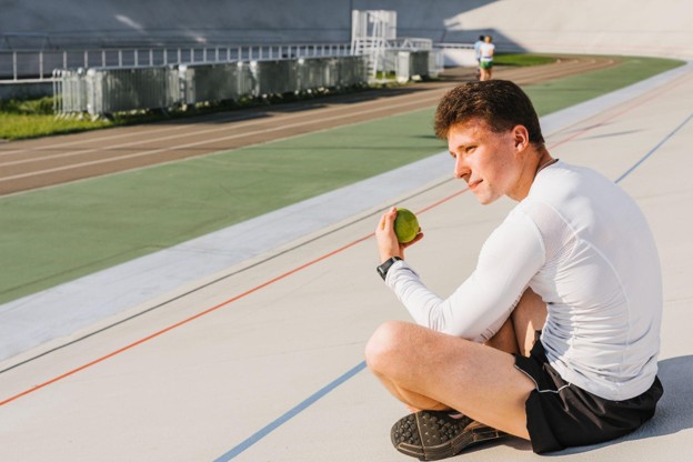 A young male athlete sitting comfortably near an outdoor track holding a tennis ball, representing a positive recovery from common types of overuse injuries.