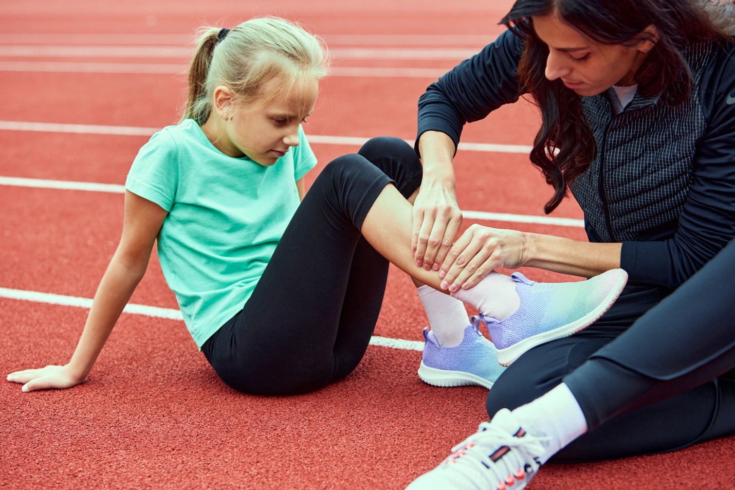 A young athlete sitting on a track with her trainer who is holding her ankle.