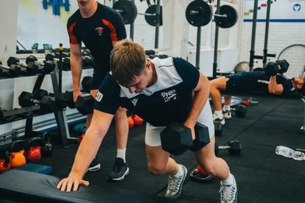 A focused athlete performing dumbbell rows in a gym as part of a strength and conditioning program.