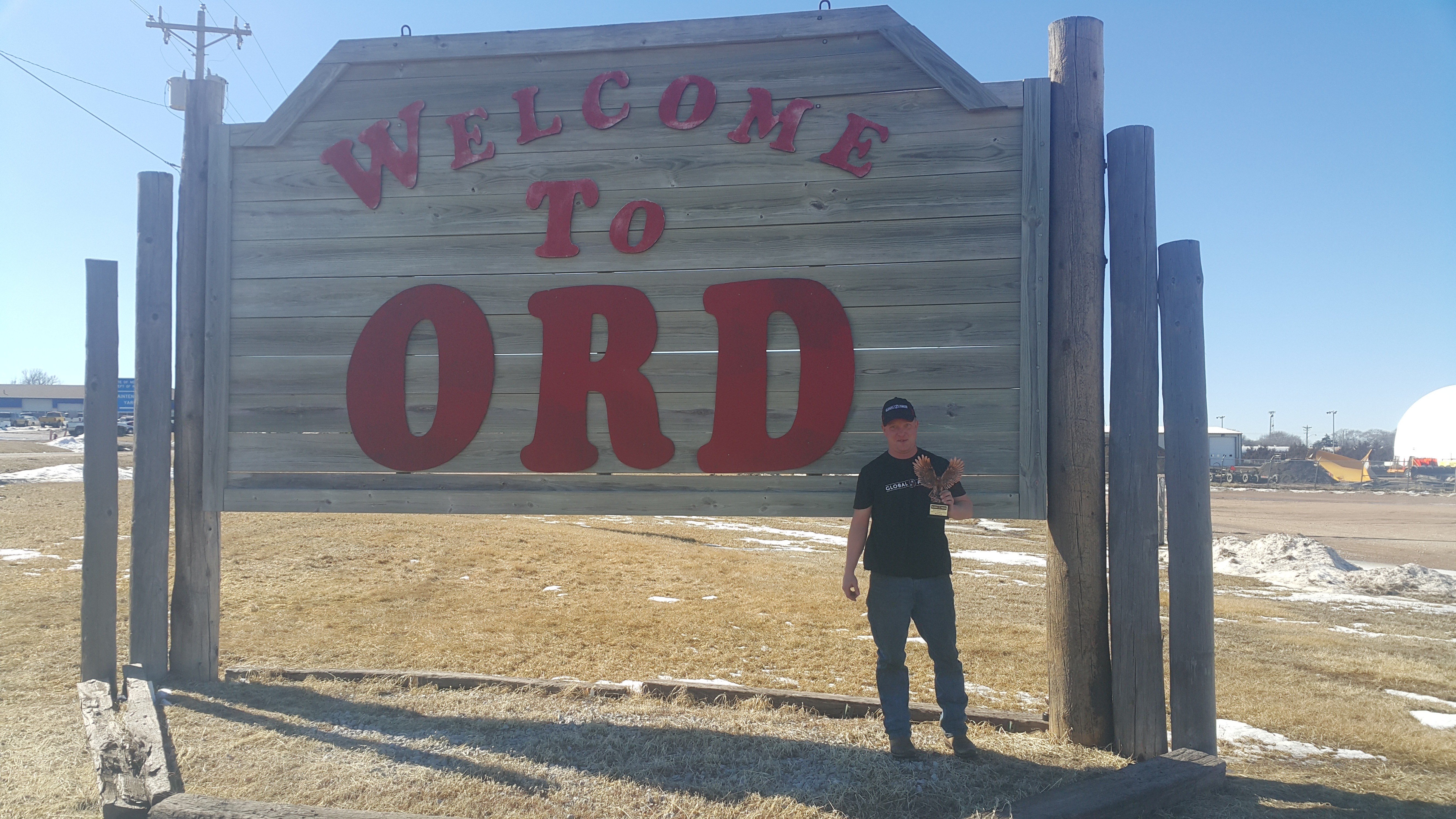 A person standing next to a large wooden sign that reads ‘WELCOME TO ORD’ in bold red letters against the wood’s natural color, with a clear blue sky in the background