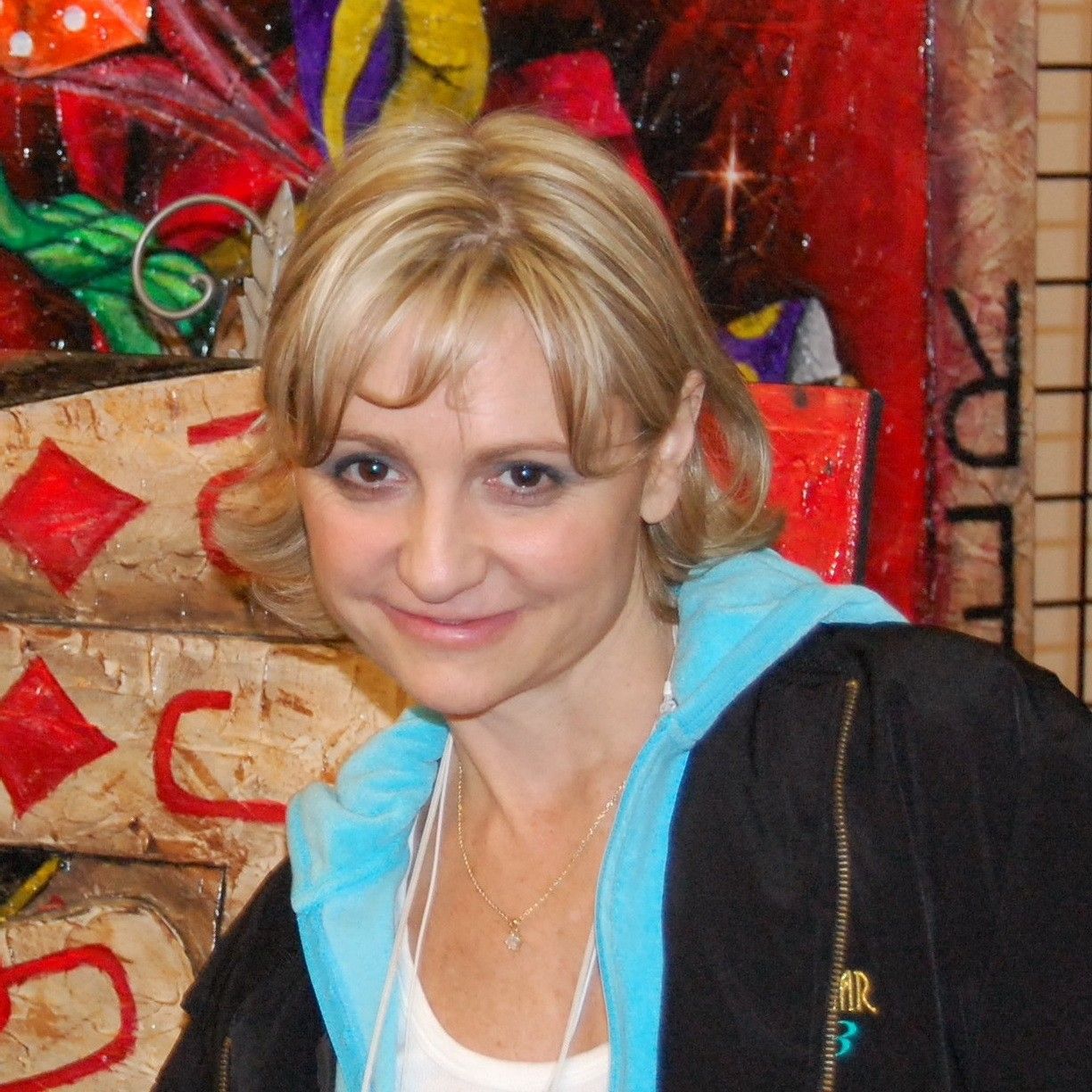 Colorful mural with abstract designs and patterns, with Jennifer Harman standing in front of it wearing a black jacket over a light blue hoodie and white top