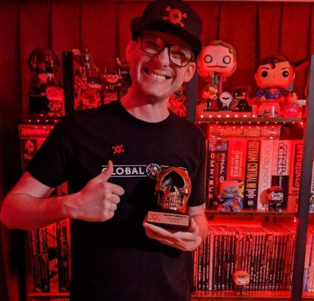 A person holding a trophy with a skull design, surrounded by collectibles and memorabilia, in a room illuminated by red lighting