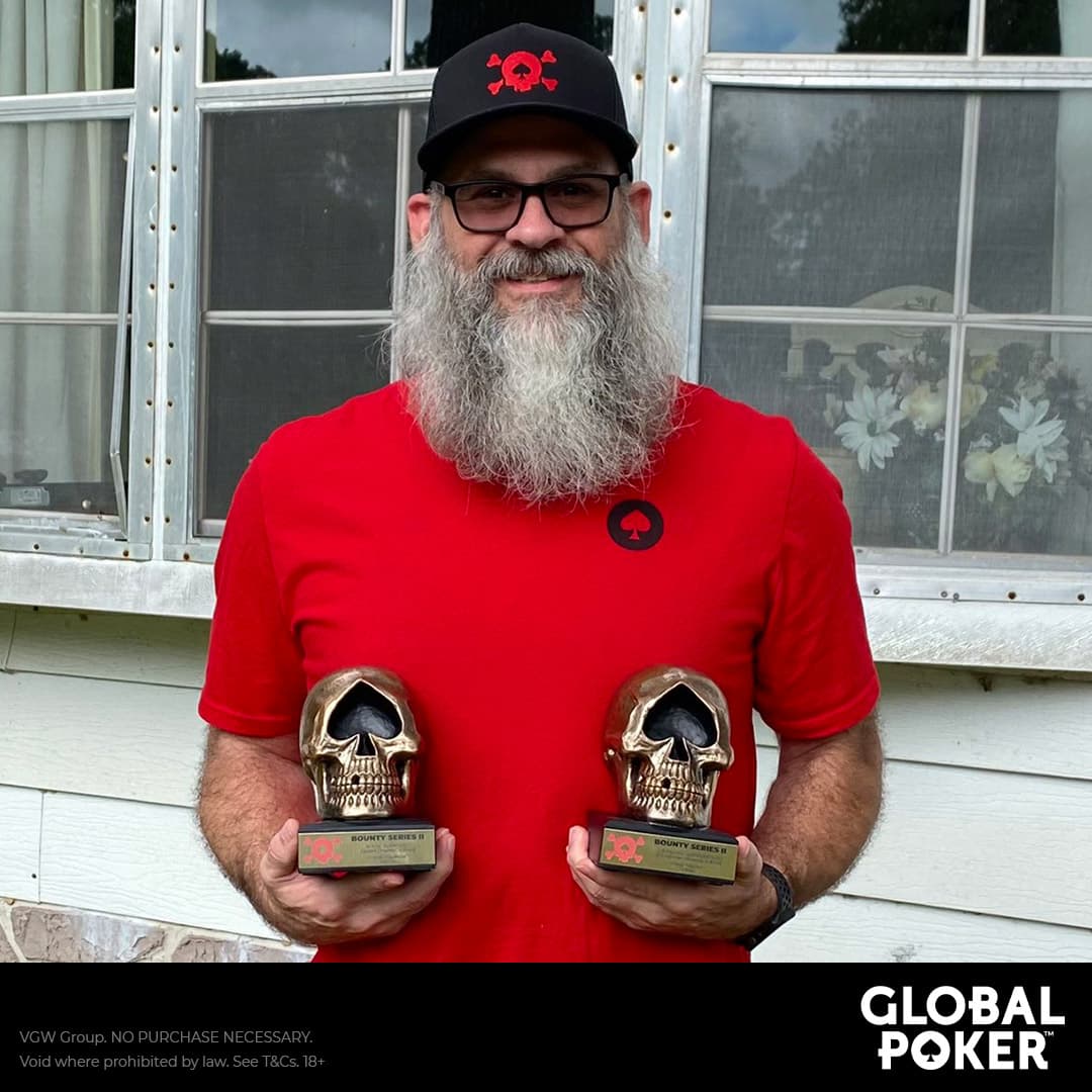 A person holding two golden skull trophies in front of a white house with windows. The person is wearing a red shirt and black cap with a red logo. The trophies are mounted on wooden bases and have inscription plates. The background shows the exterior of a house with white walls and multiple windows; curtains with floral patterns can be seen through the windows