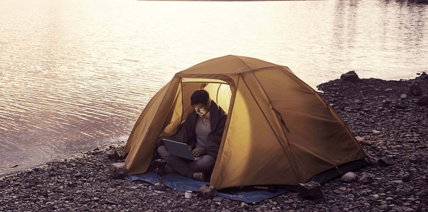 A person working on a laptop inside a yellow tent by a serene lake during sunset