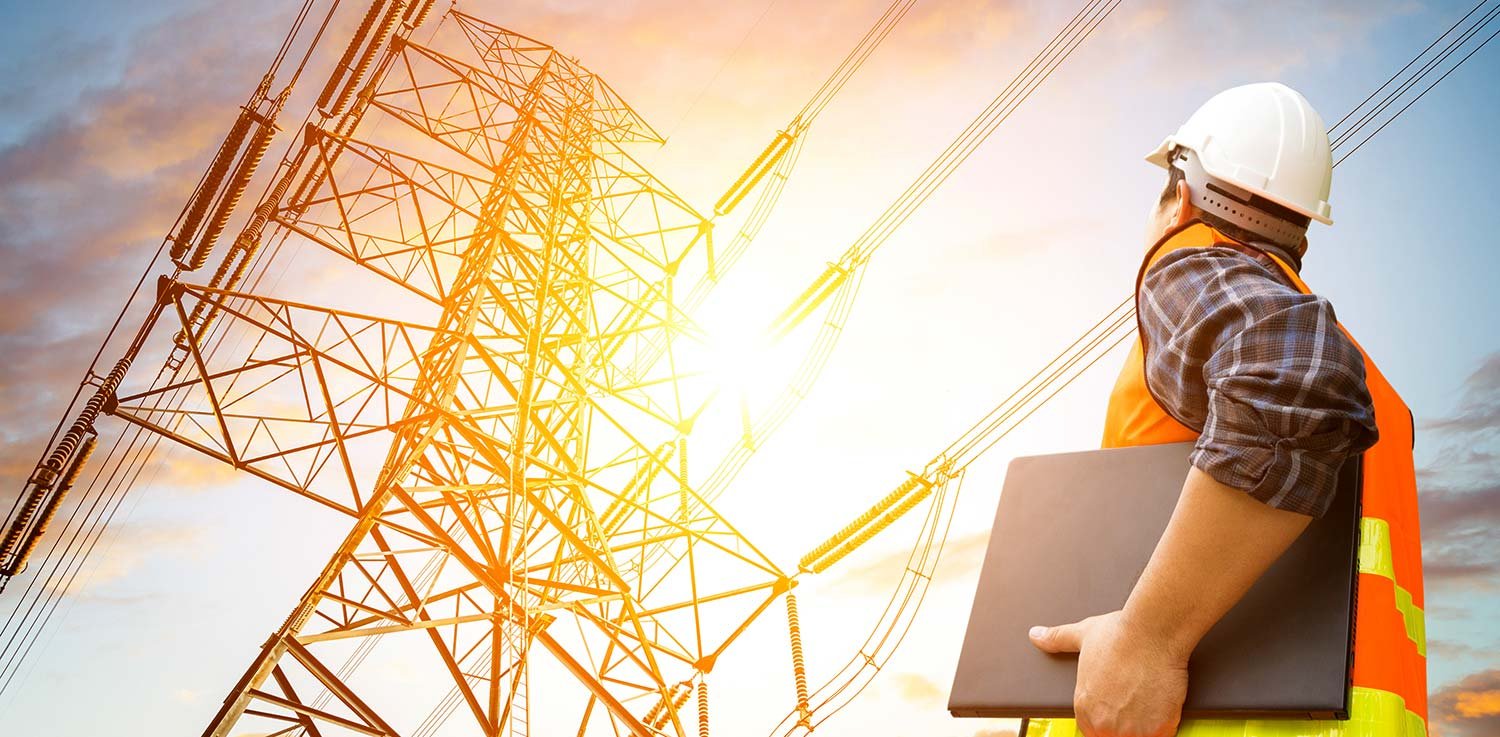 Man from local electric company in hard hat carrying computer and looking up at power lines