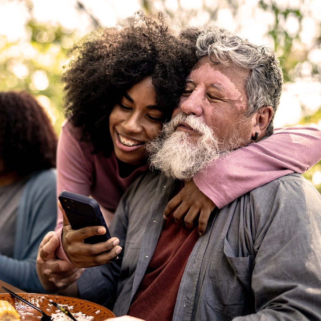 Young woman hugs an elderly family member warmly as they look at her smartphone together.
