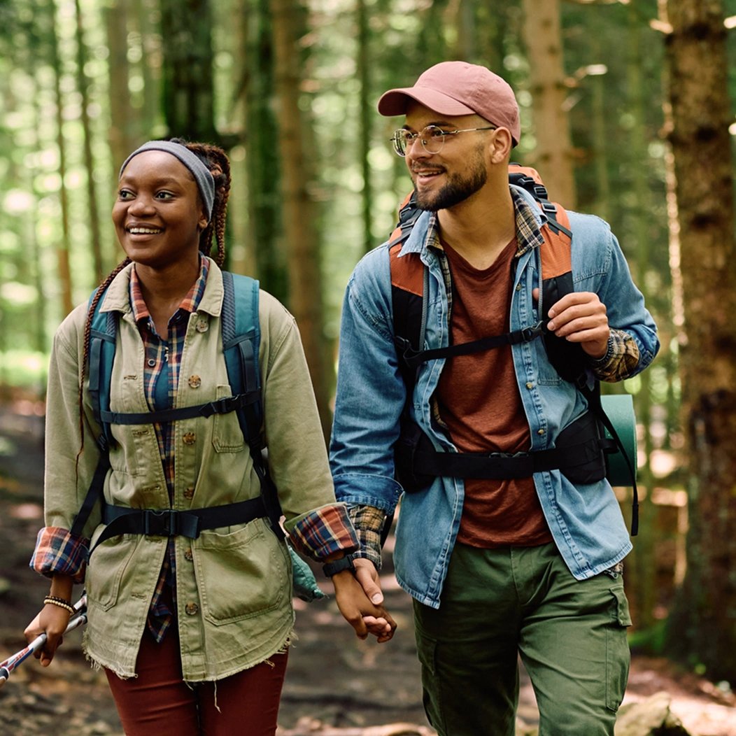 A young couple wearing outdoor clothing and backpacks hikes confidently through the woods thanks to their affordable life insurance coverage.