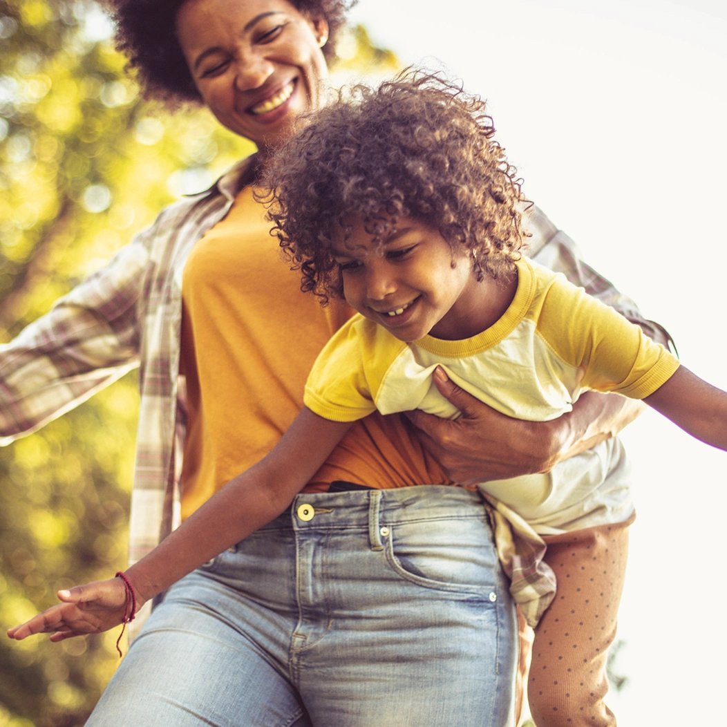 A smiling mom carries her young son as he smiles and pretends to fly.