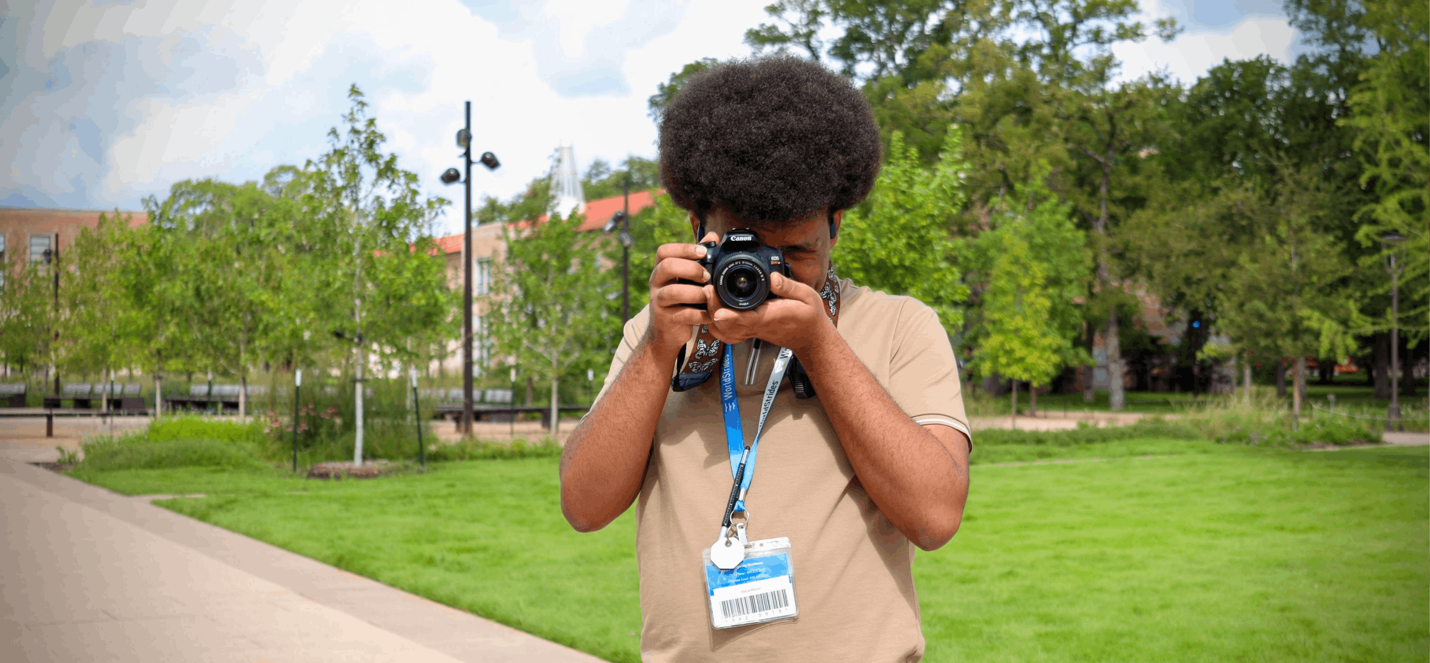 A person with an ID badge takes a photo with a camera outdoors on a sidewalk next to a grassy area and trees.