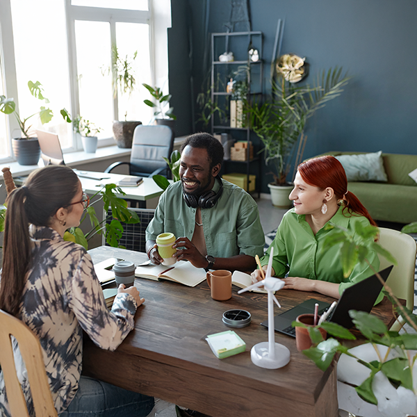 Three people sitting around a table in a modern, plant‑filled workspace, talking together with notebooks, coffee cups, and a laptop on the table.