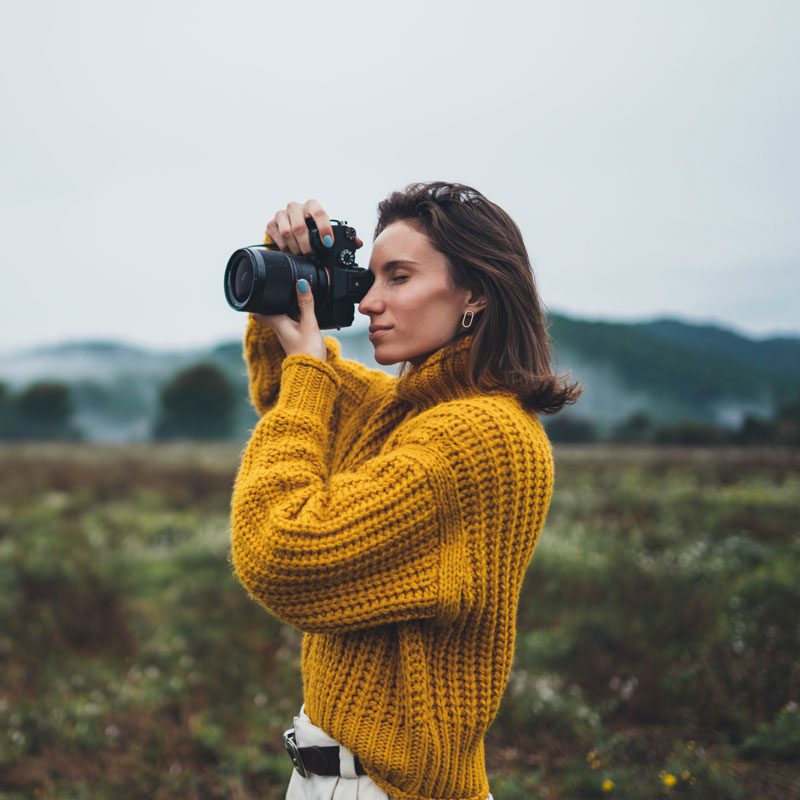 woman in a yellow jumper standing outdoors holding a camera 