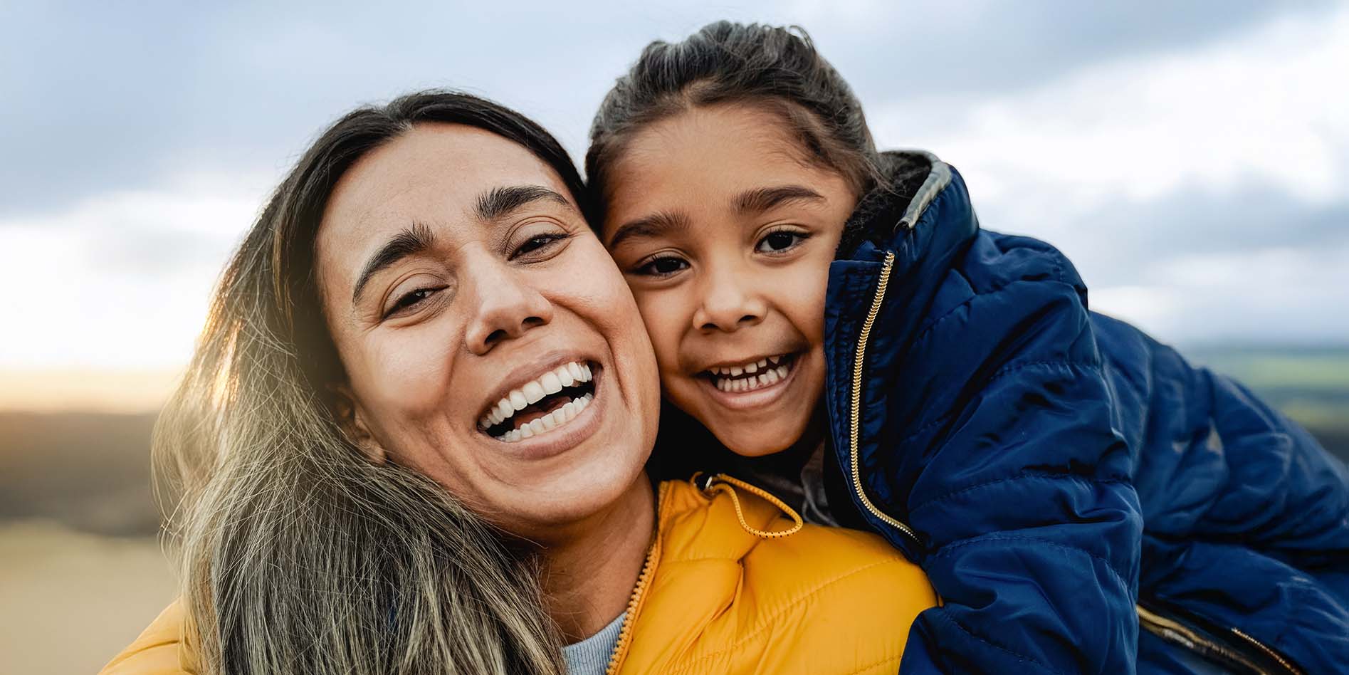An adult and a child outdoors wearing warm puffer jackets, with the child leaning close against the adult under a cloudy sky.