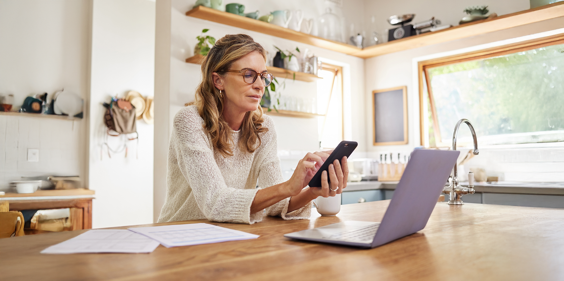 Woman wearing glasses reviewing financial information on her phone with a laptop and paperwork on a kitchen bench at home, planning for end of financial year.