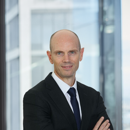 Professional head‑and‑shoulders portrait of a person wearing a business suit and tie, standing with arms crossed in front of a modern office building interior with glass walls.