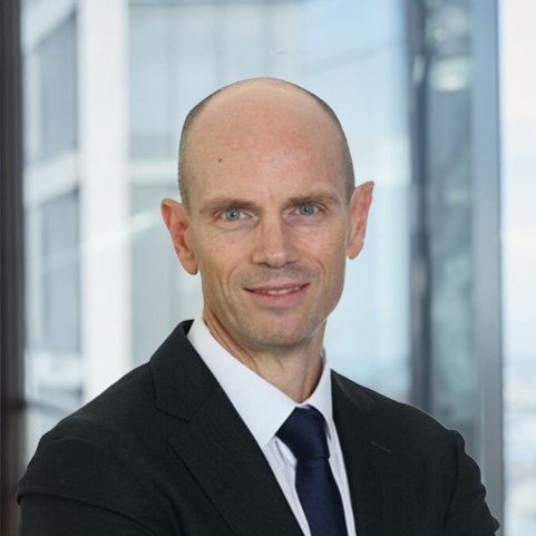 Professional head‑and‑shoulders portrait of a person wearing a business suit and tie, standing with arms crossed in front of a modern office building interior with glass walls.