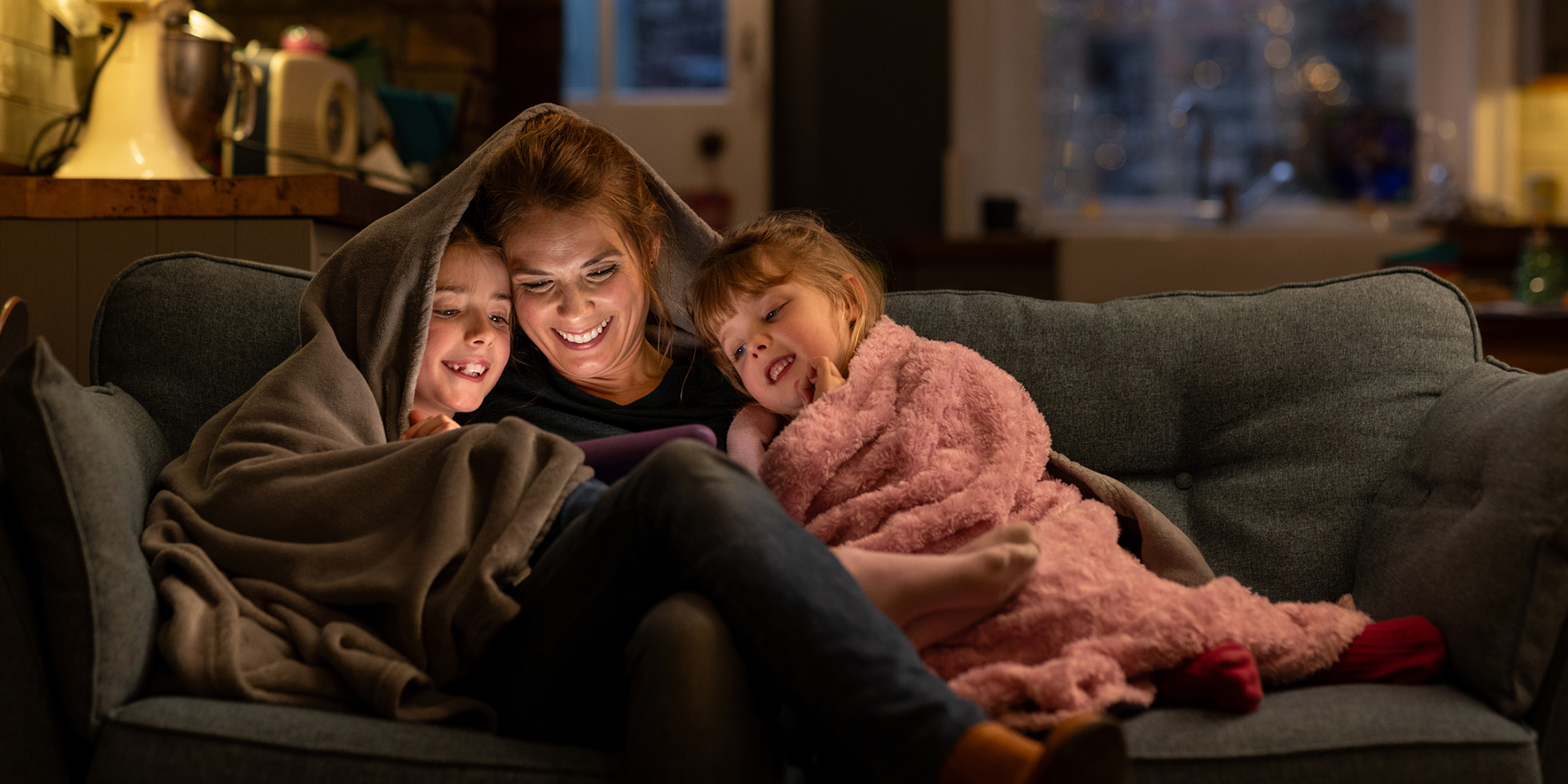 woman-and-two-todler-girls-looking-at-tablet-under-blanket