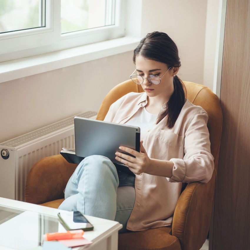 2CABJWY Konzentrierte junge Frau mit Brille, die im Sessel sitzend am Laptop arbeitet