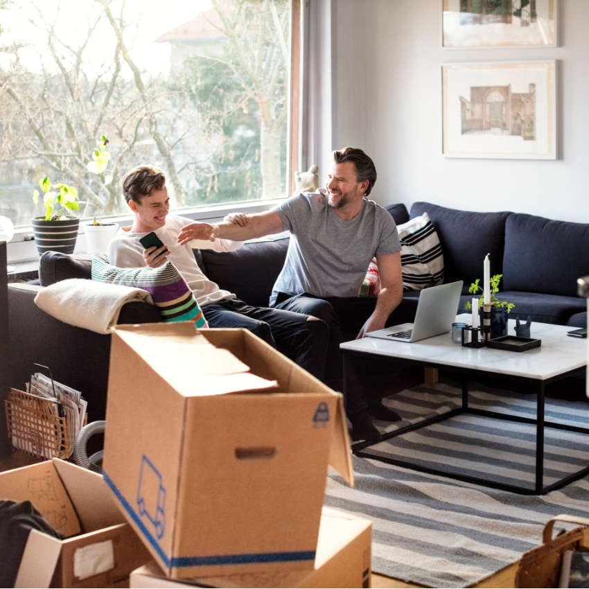 Vater und Sohn, die zusammen in einem Wohnzimmer mit Umzugskartons auf einem Sofa sitzen und lachen