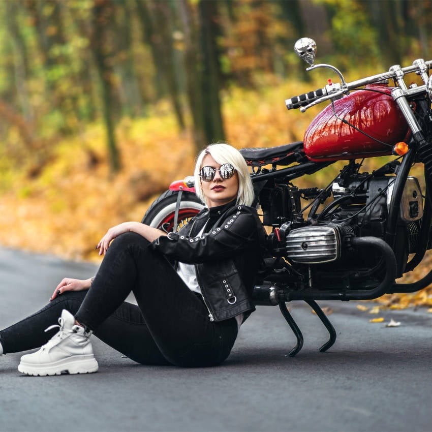 2CWCD2E Frau mit Sonnenbrille sitzt vor einem roten Motorrad auf einer Straße im herbstlichen Wald