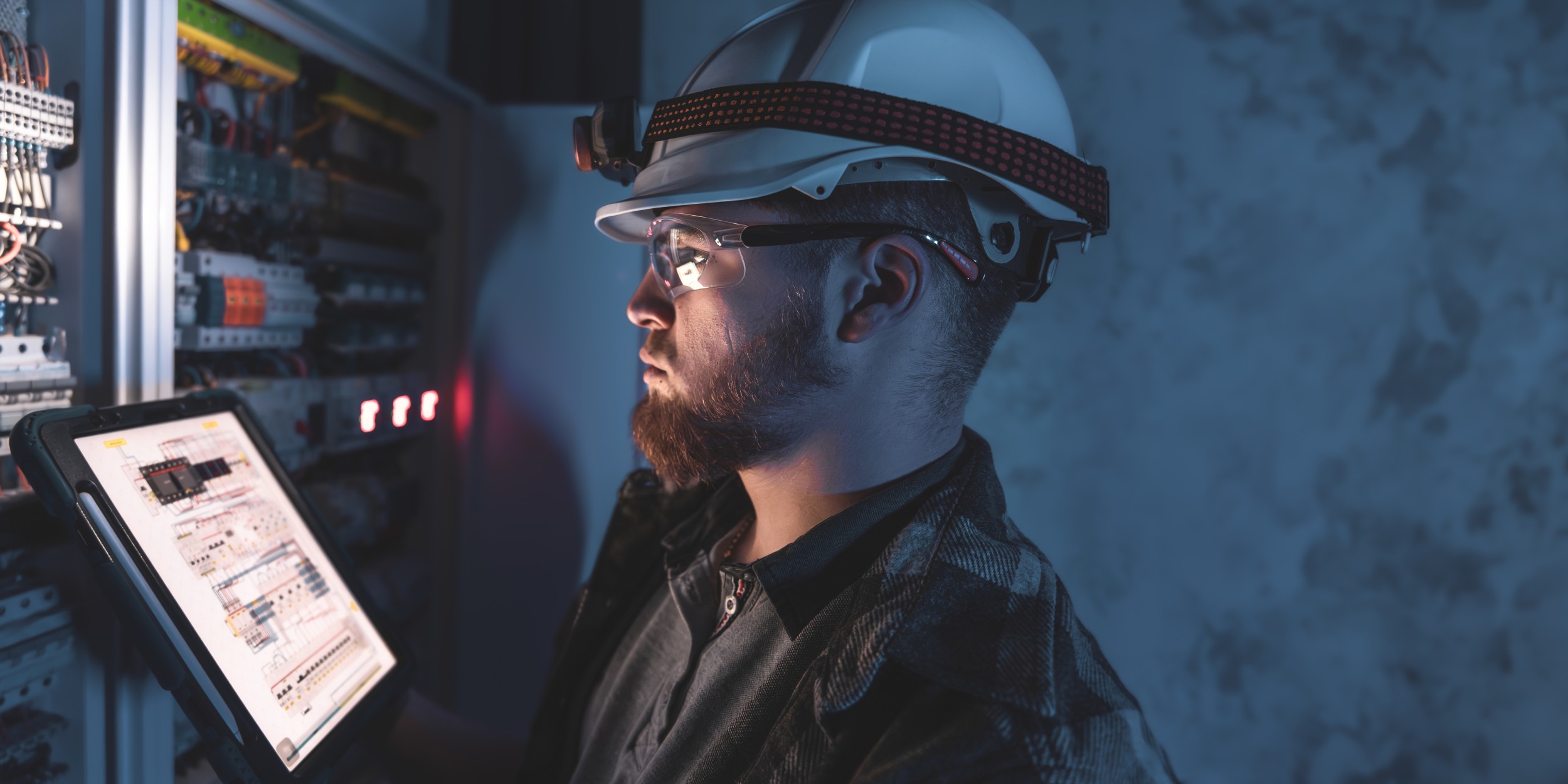 An engineer wearing a hard hat and headlamp inspects an open electrical control panel while reviewing circuit diagrams on a tablet, in a dimly lit industrial setting.