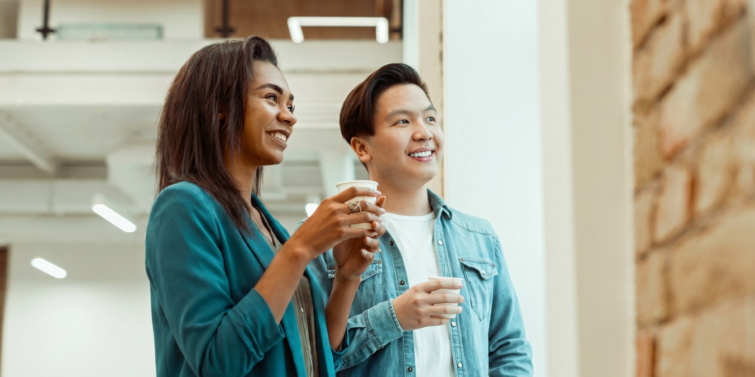 two colleagues holding coffee in office hallway