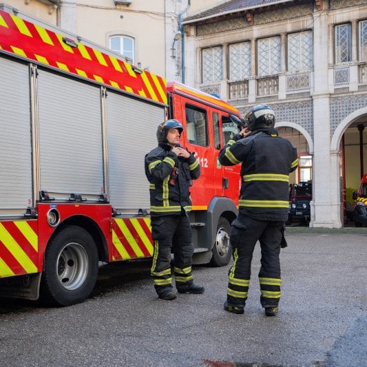  Two firefighters in uniform stand next to a red fire truck in a courtyard, with another emergency vehicle visible in the background.