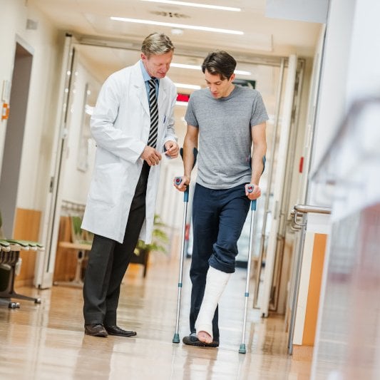A doctor walks beside a patient using crutches with a leg cast in a hospital corridor.
