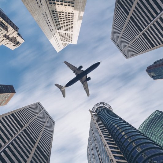 Airplane flying over tall city skyscrapers, viewed from below against a blue sky.