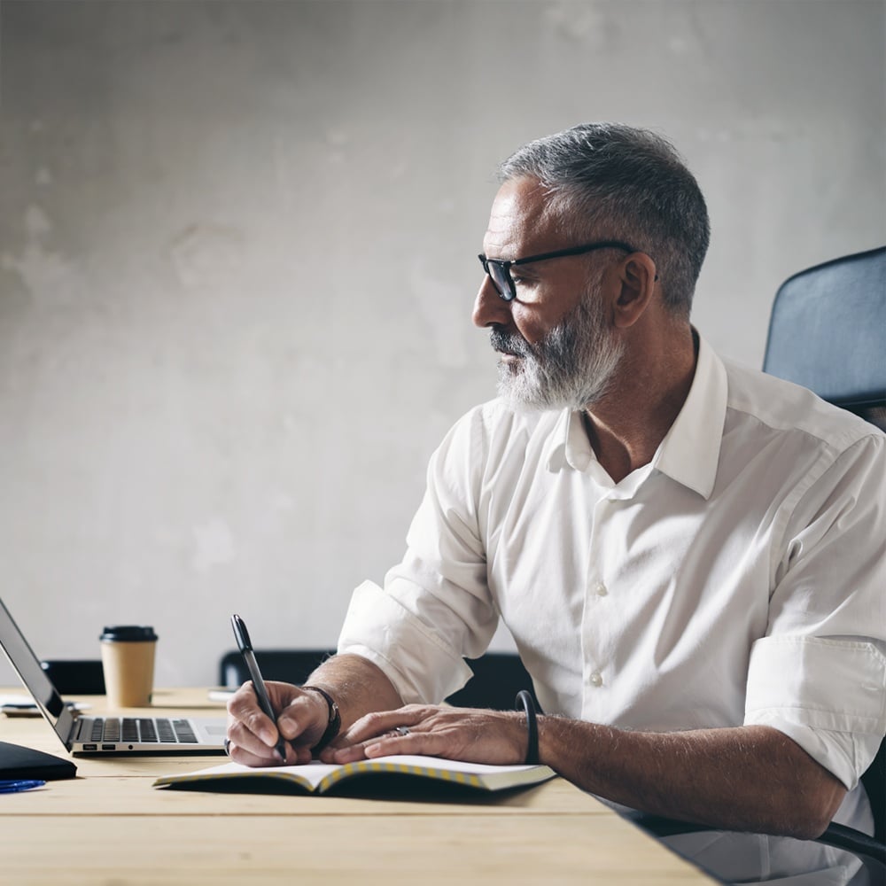 Man writing in notebook at desk with laptop and coffee.
