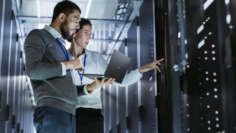 Male it specialist holds laptop and discusses work with female server technician. they're standing in data center, rack server cabinet is open.