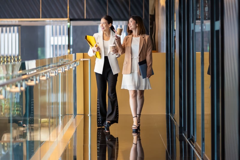 Two asian businesswomen walking and talking during coffee break in modern office or coworking space.