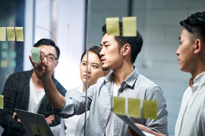 Young  Asian entrepreneur of small company putting a adhesive note on glass in office during team meeting formulating business strategies.