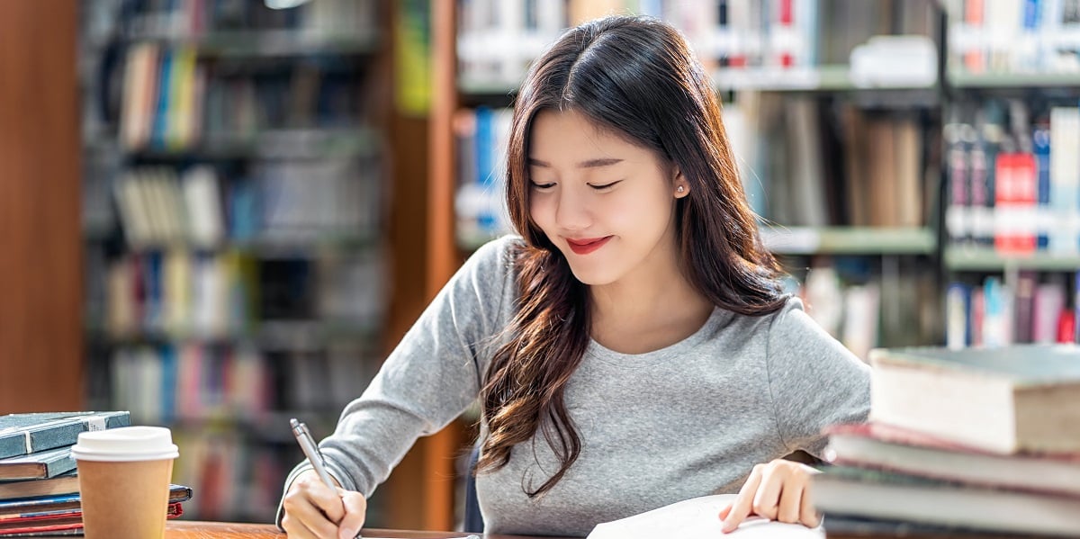 Asian female student writing notes in a library next to a pile of books.