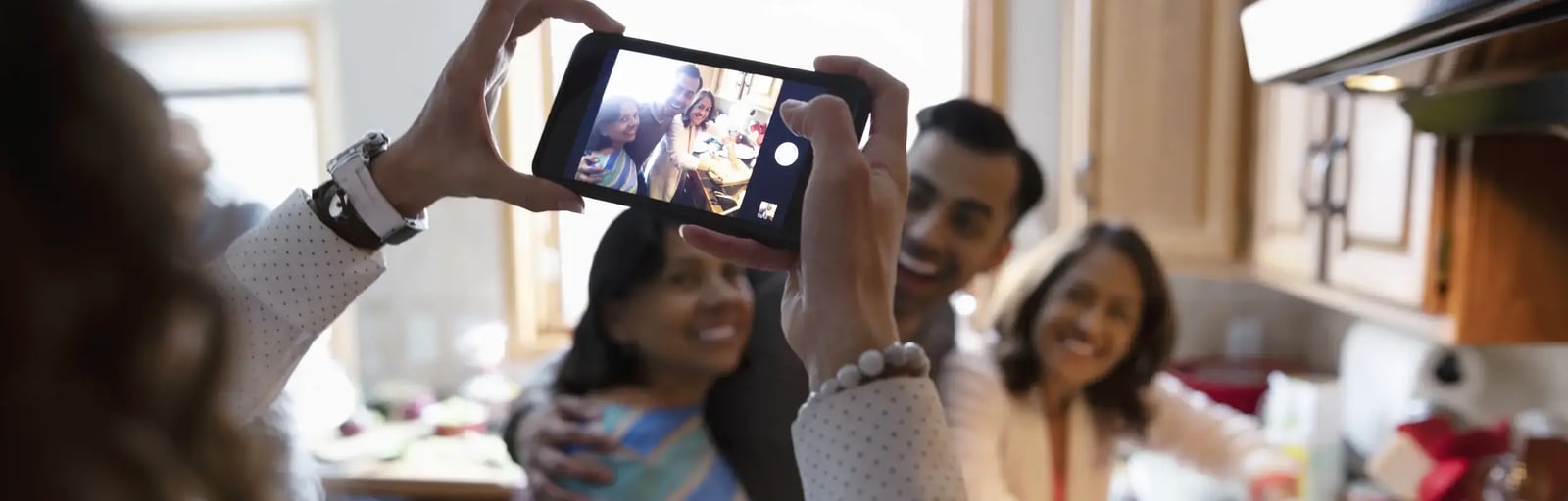 A woman is holding her phone taking a picture of a family group preparing a meal in the kitchen