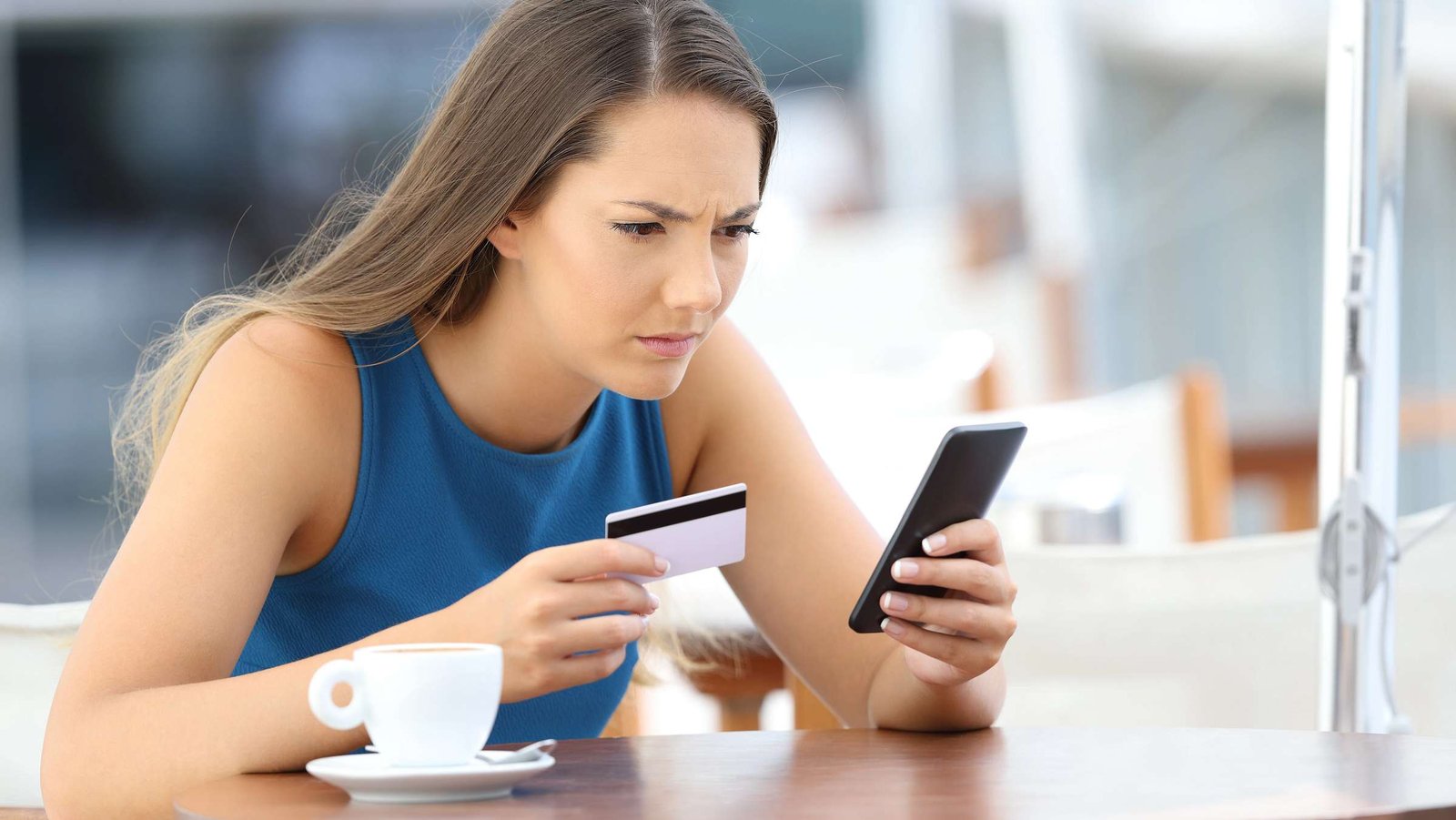 Eine Dame sitzt im Cafe, hat die Kreditkarte und das Handy in der Hand und schaut entgeistert.
