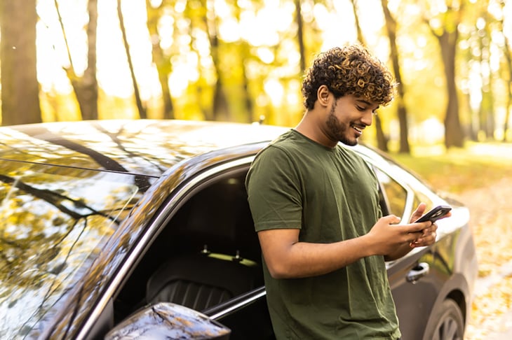 Junger Mann steht mit Handy in der Hand neben dem Auto