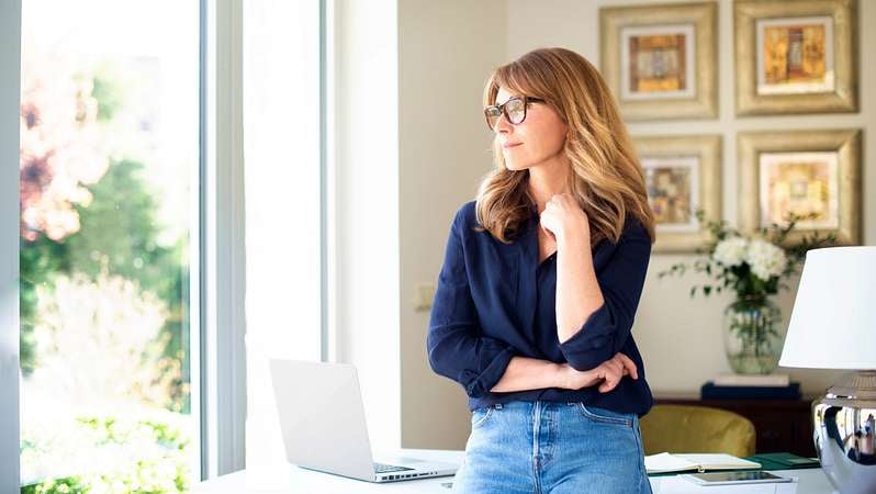 Junge Dame blickt nachdenklich aus dem Fenster und steht vor dem geöffneten Laptop.