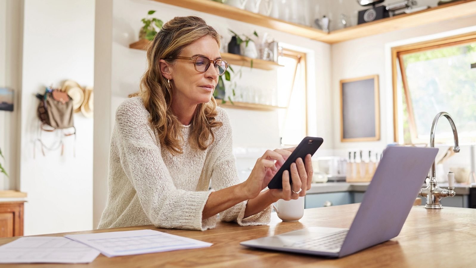 Dame sitzt mit Handy vorm Laptop und rechnet ihre Pension aus.