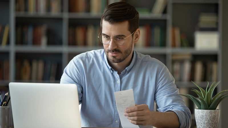 Mann sitzt mit Rechnung vorm Laptop.